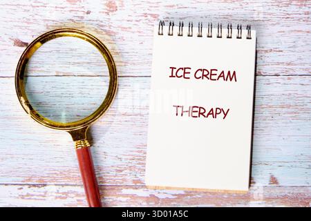 A notepad with the words ice cream therapy sits beside a magnifying glass on a rustic wooden table. Stock Photo