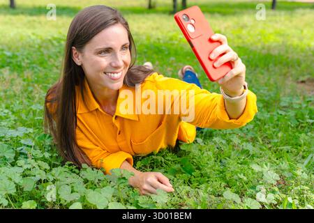 Smiling woman in yellow shirt taking selfie with smartphone lying on green grass Stock Photo