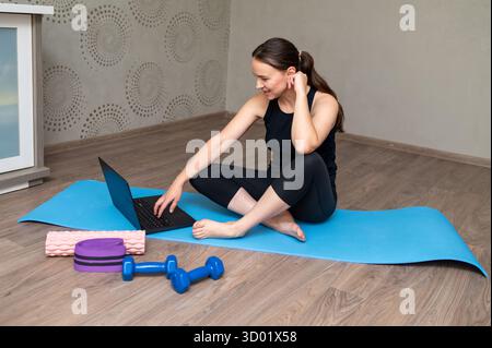 Smiling woman in sportswear sits on yoga mat with laptop and dumbbells, doing home workout and online fitness training concept Stock Photo