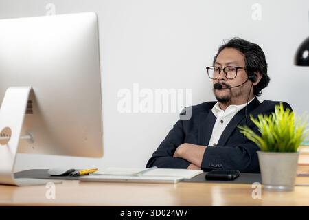 A tired young man wearing headphones falls asleep while attending an online meeting, representing remote work fatigue, stress, and modern work-from-ho Stock Photo