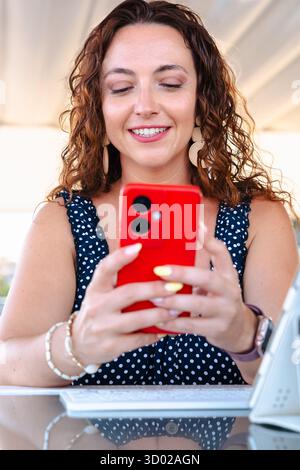 Elegant woman using red smartphone at table with drink and tablet outdoors Stock Photo