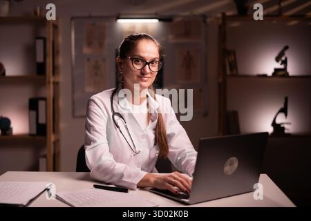 female doctor in eyeglasses smiling looking at camera while working with laptop on medical consultation working late night in hospital. copy space Stock Photo