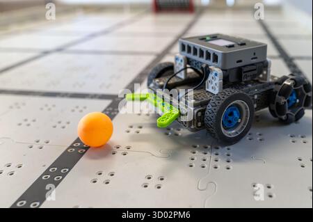 Small robot car moves toward a vibrant orange ball on a patterned floor in a tech environment. Stock Photo