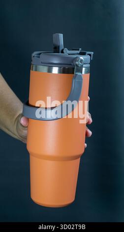 Orange tumbler bottle with open straw lid held in hand, portrait orientation on black background, reusable drinkware shown in active daily use style Stock Photo