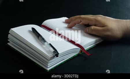 Side view of notebook and pen on book with male hand above, placed on black table against black background, minimal dark-themed writing concept Stock Photo