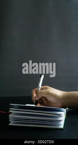 Male hand holding notebook and pen in side view, portrait orientation on black table with black backdrop, ready-to-write dark composition style Stock Photo