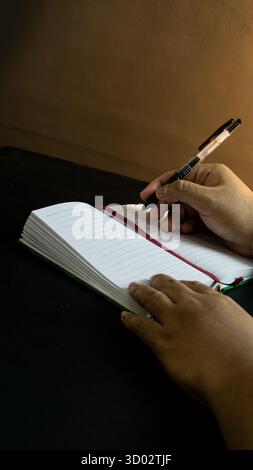 Portrait side view of male hand with notebook and pen under warm light, placed on black table with black backdrop, dark minimal composition Stock Photo
