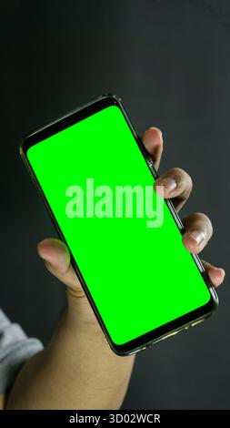 Left hand gripping smartphone angled from the side, green screen on display, isolated against dark background for digital or tech presentation Stock Photo