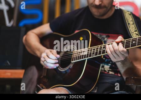 A young Caucasian man with a beard plays an acoustic guitar. He wears a black t-shirt and is focused on his music in a casual setting. Stock Photo
