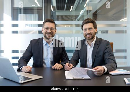 Two smiling adult businessmen signing a legal document, finishing a successful deal and agreement, representing collaboration and cooperation in modern office Stock Photo