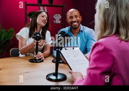 Three diverse individuals, a host and a couple guests, smiling and engaging in conversation while recording an audio podcast or live broadcast with mi Stock Photo