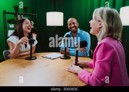 Three diverse adult hosts, a man and two women, sitting around a table with microphones, engaging in a lively podcast discussion or interview in a cur Stock Photo