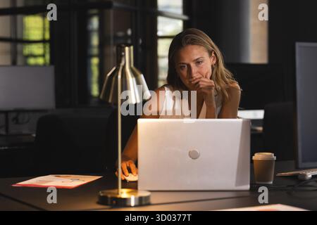 Focused woman working late on laptop in modern office with coffee. Business, technology, concentration, productivity, deadline, workspace || Model released Stock Photo