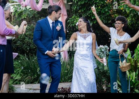 Bride and groom walking joyfully as guests celebrate with bubbles outdoors. Wedding, celebration, marriage, happiness, festive, ceremony || Model released Stock Photo