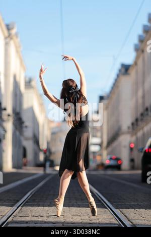 Ballet dancer in a black dress performs an elegant en pointe pose on cobblestone tram tracks, framed by classic buildings and blue sky. || Model released Stock Photo
