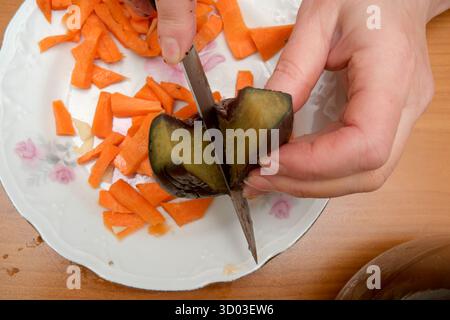 A person skillfully slices an eggplant while preparing carrots in a warm kitchen. Stock Photo