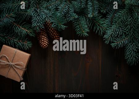 A small rustic gift box sits below a border of dark blue spruce branches and pine cones on a wooden background. Stock Photo