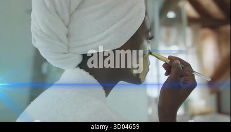 Woman applying golden facial mask with makeup brush in bathroom, wearing bathrobe and towel-turban Stock Photo