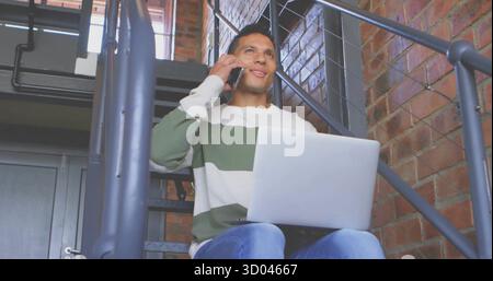 Talking man holding smartphone and using laptop on lap at loft workspace, with metal staircase Stock Photo