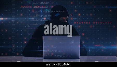 Typing hacker in balaclava and hoodie on laptop at desk in dark room, code, error messages Stock Photo