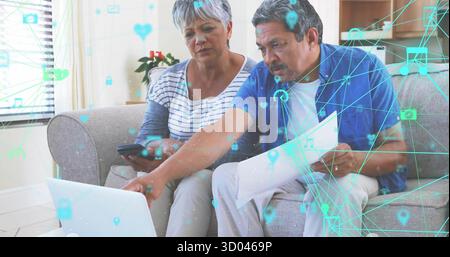 Sitting senior couple focusing on laptop in living room with smartphone, documents, network icons Stock Photo
