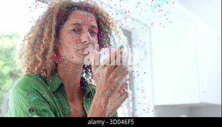 Standing curly haired woman wearing green shirt in bright kitchen by window, holding white mug Stock Photo