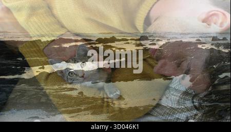 Holding man wearing mustard sweater cradling gray and white cat on bed, with coastal overlay Stock Photo