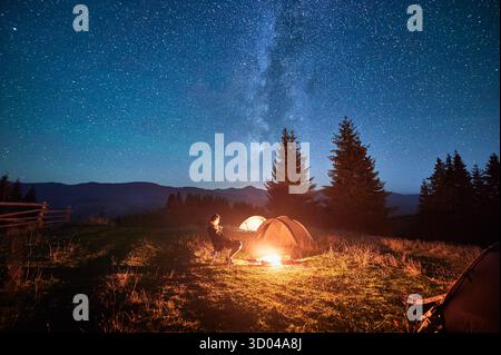 Hiker woman relaxes by campfire, illuminated by its warm glow, with tent nearby under stunning, star-filled sky. Milky Way stretches overhead, adding magical touch to serene mountain setting. Stock Photo