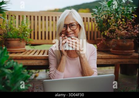 Older woman having cup of tee while working from homeoffice, sitting on terrace. Stock Photo