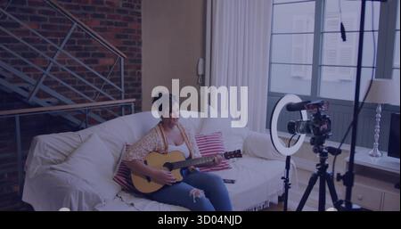 Strumming woman wearing ripped jeans on sofa in living room, with acoustic guitar and camera setup Stock Photo