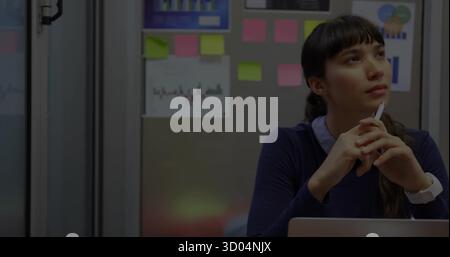 Holding pen, woman in navy sweater gazing upward in office booth with open laptop, copy space Stock Photo