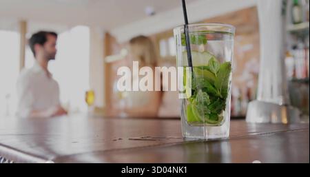 Showcasing mojito glass with mint, cucumber slices on bar counter, with blurred couple chatting Stock Photo