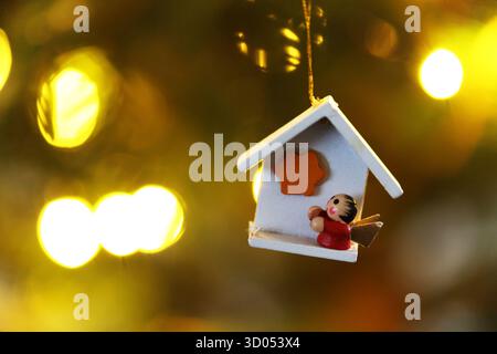 Miniature white wooden house ornament with a person figure inside hanging on a Christmas tree with warm bokeh lights in the background Stock Photo