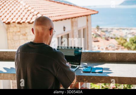 Freelancer bald fency man enjoying remote work on sunny terrace surrounded by greenery with sea view. Digital nomad using laptop embracing freedom fle Stock Photo