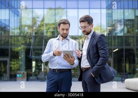 Two professional businessmen engaging in a serious discussion while viewing information on a digital tablet outdoors in front of a modern glass office building Stock Photo