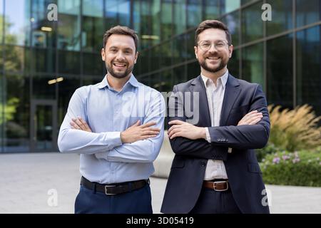 Two smiling adult businessmen, one with glasses, standing confidently with crossed arms outside a modern office building, representing teamwork and success Stock Photo