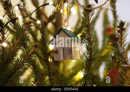 Wooden house ornament hanging on a Christmas tree, surrounded by fir branches and warm fairylights in the background Stock Photo