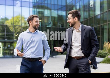 Two smiling adult businessmen walking and having a conversation outside a modern corporate office building, discussing business strategy or partnership Stock Photo