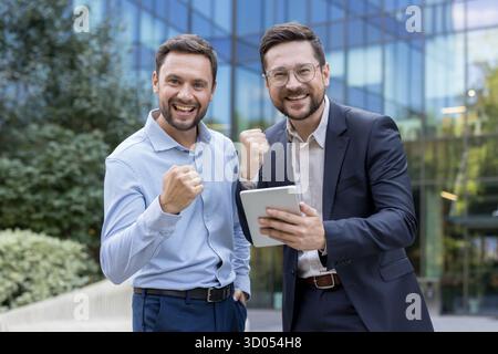 Two happy businessmen celebrating an achievement, clenching fists and smiling at camera while one is holding a digital tablet with a modern office building in background Stock Photo