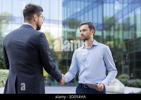 Two professional businessmen shaking hands outside a modern office building, symbolizing agreement, successful negotiation, and a new partnership forming between colleagues during a business meeting Stock Photo