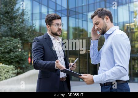 Two businessmen having an intense argument and conflict outdoors, one appearing stressed and covering his head while holding documents, the other gesturing as he speaks Stock Photo