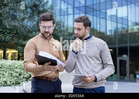 Two businessmen colleagues discussing important financial documents and corporate data, analyzing figures and planning strategies outdoors in front of a modern office building Stock Photo