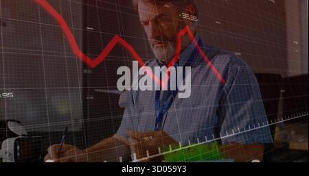 Writing senior male analyst taking notes in notebook at office desk, with financial data overlay Stock Photo