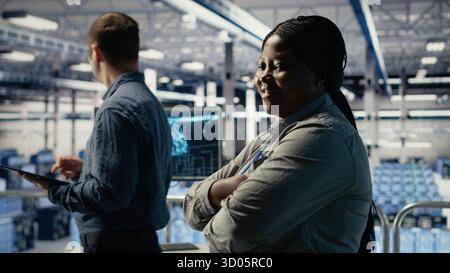 Portrait of smiling data center manager working to improve AI rigs processing speed and efficiency. Happy team leader in server hub manages gear to support artificial intelligence workloads, camera B Stock Photo