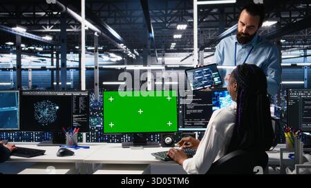 Female programmer talking with manager in data center, reviews AI diagnostics on green screen PC. IT expert and supervisor optimizes artificial intelligence systems using mockup computer, camera A Stock Photo