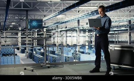 Data center admin on industrial platform checks AI dashboard using laptop. Man in server farm tests artificial intelligence tools on notebook to ensure reliable performance, camera A Stock Photo