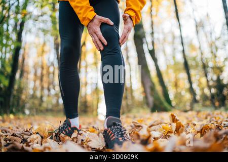 Close-up image of runner athlete dressed sporty on the running path standing and feeling acute knee pain while morning jogging in yellow autumn park. Stock Photo