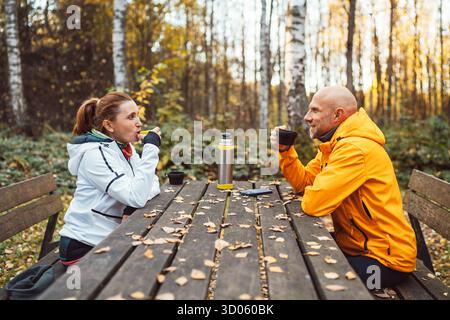 Middle-aged couple resting after walk enjoying hot tea from thermos at wooden picnic table in autumn forest. Active lifestyle, outdoor relaxation cozy Stock Photo