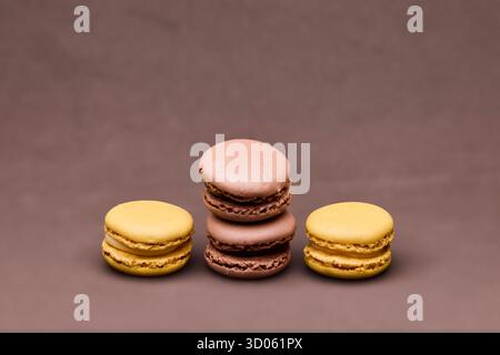 Three macarons in chocolate and yellow tones arranged on brown background in studio lighting. The minimal setup highlights texture and color contrast Stock Photo