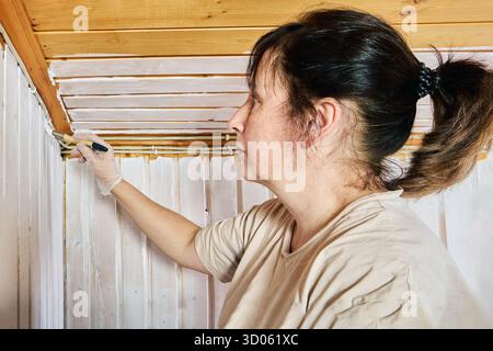 Decorating job, female worker covering timber cladding with white paint brush strokes transforming natural boards into bright finish. Stock Photo